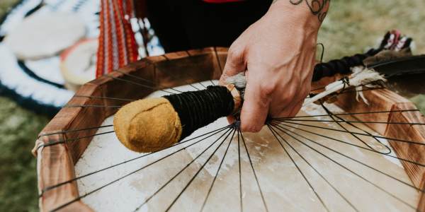 Close-up of a hand holding a drumstick over a traditional drum with strings, featuring intricate tattoos on the arm.