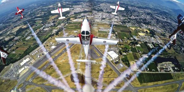 Five red-and-white aerobatic aircraft fly in formation above an airport and surrounding farmland, leaving white smoke trails that fan out beneath the planes.